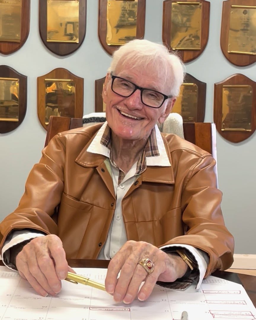 Jerry Foster smiling at a desk with award plaques on the wall behind him